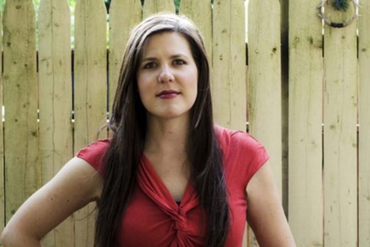 Woman with long dark hair wearing a red top standing in front of a wooden fence