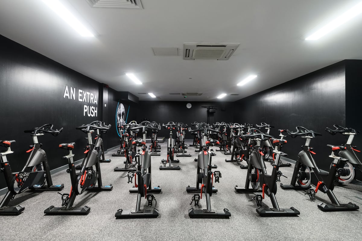 PureGym Newcastle Longbenton: Rows of stationary exercise bikes in a dimly lit studio with black walls and neon signage.