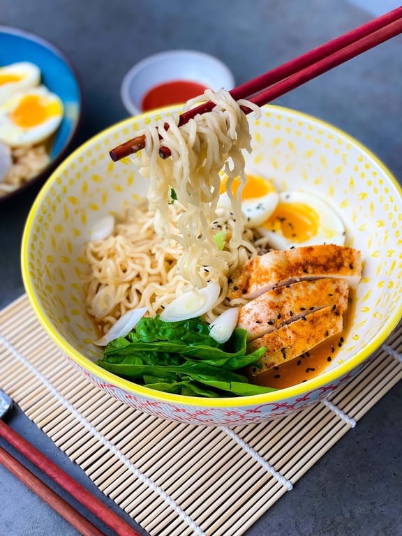 Chopsticks lifting noodles from a bowl with chicken, spinach, boiled egg, and broth.