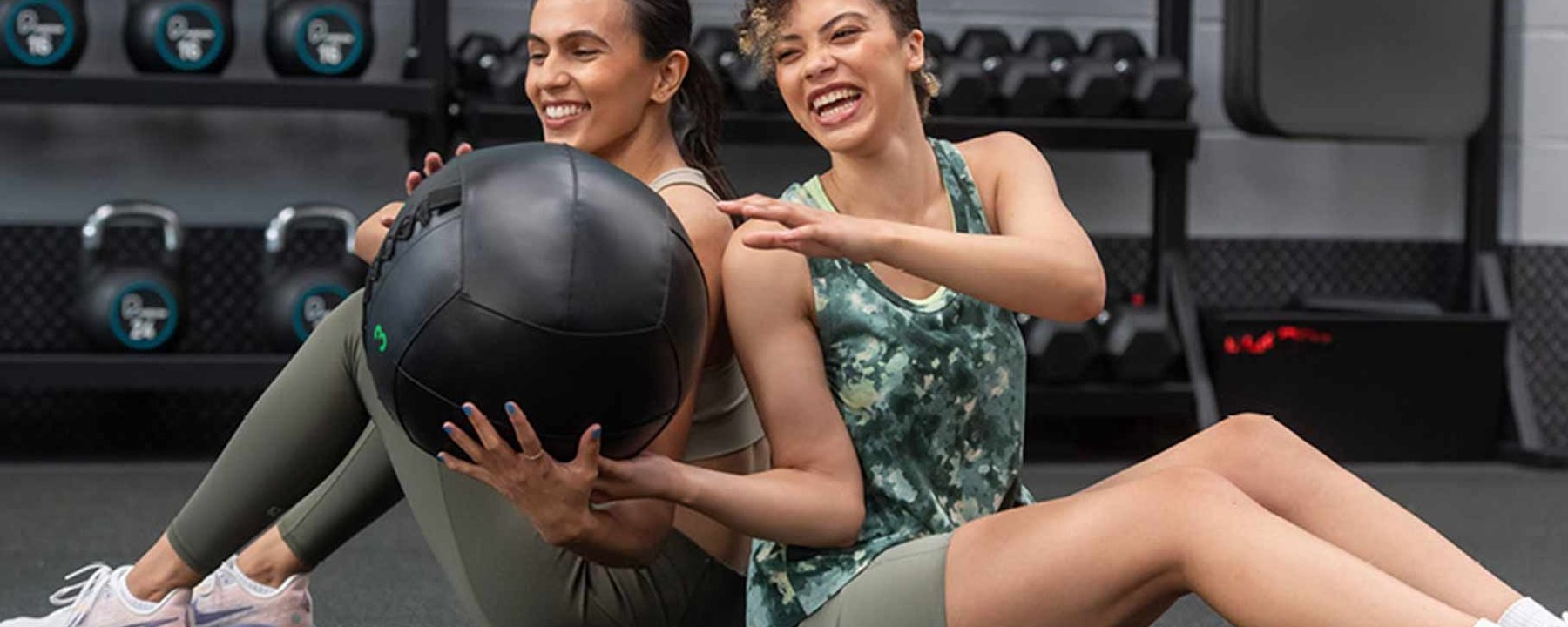 Deux femmes assises dos à dos se passent un ballon médicinal en salle de sport