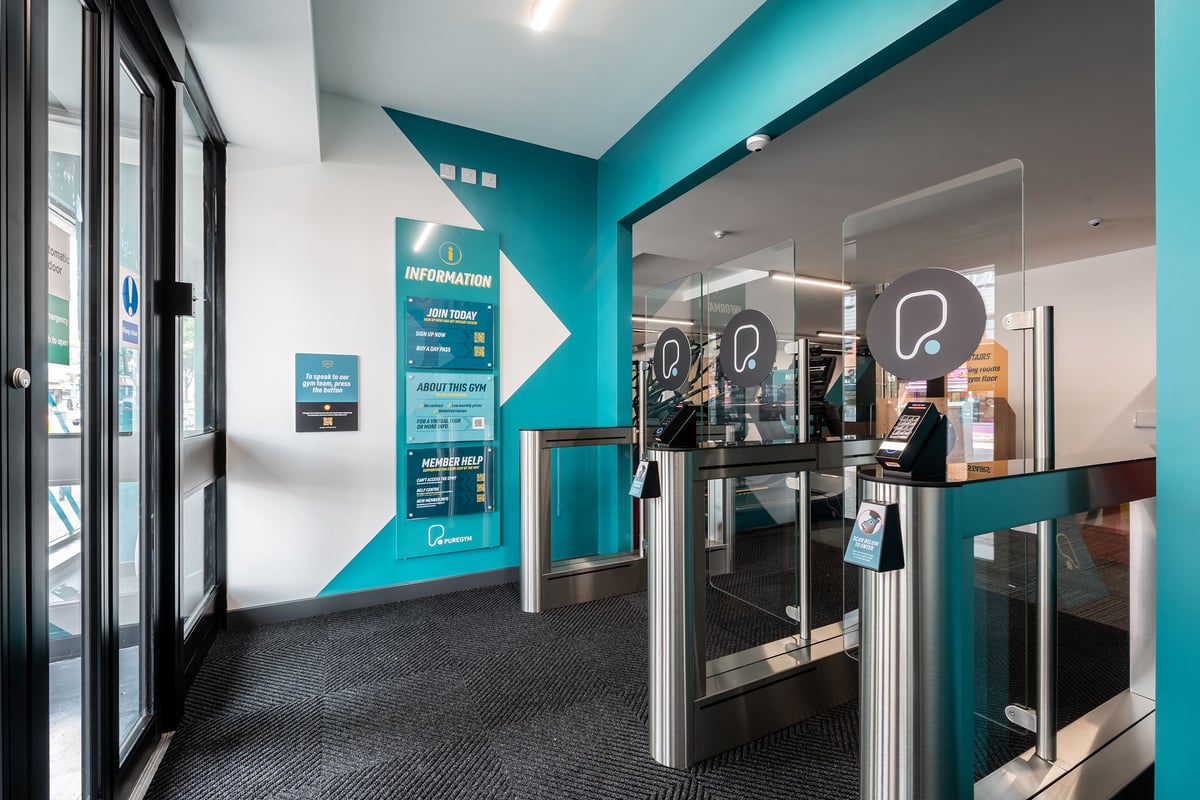 PureGym London Welling: Turnstiles, teal coloured partitions, and glass entrance doors to a gym facility.