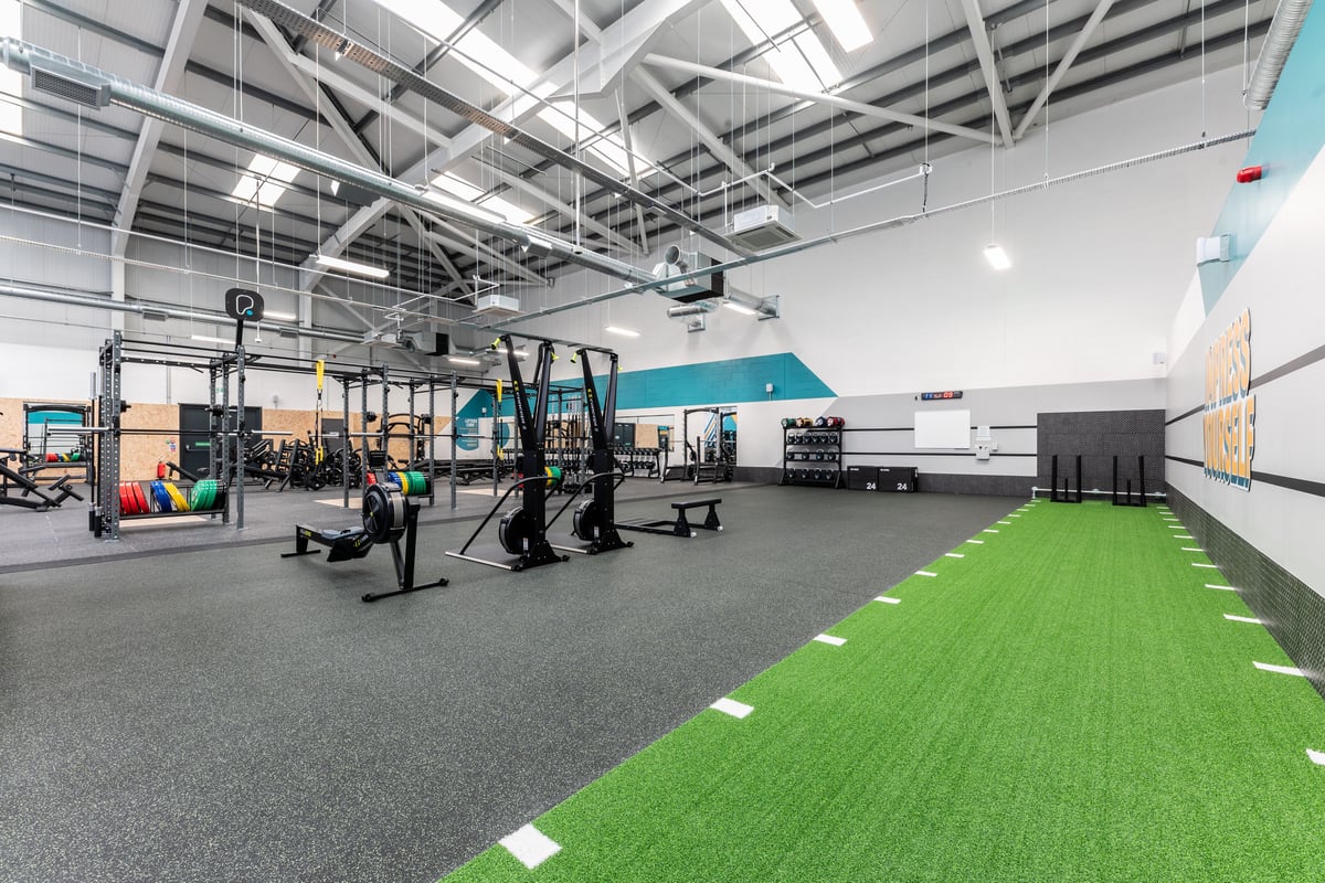PureGym Port Talbot: Gym studio with weightlifting rigs, rubberized flooring, green turf area, and mirrors along walls.