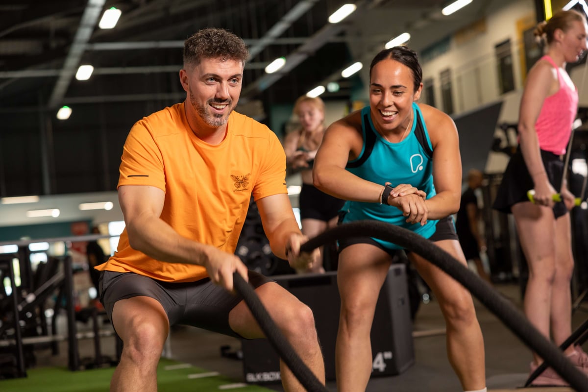 Man in orange shirt using battle ropes while woman in blue tank top encourages him in gym