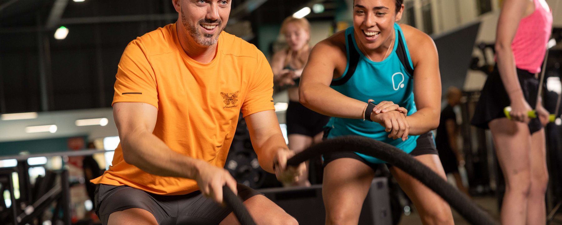 Man in orange shirt using battle ropes while woman in blue tank top encourages him in gym