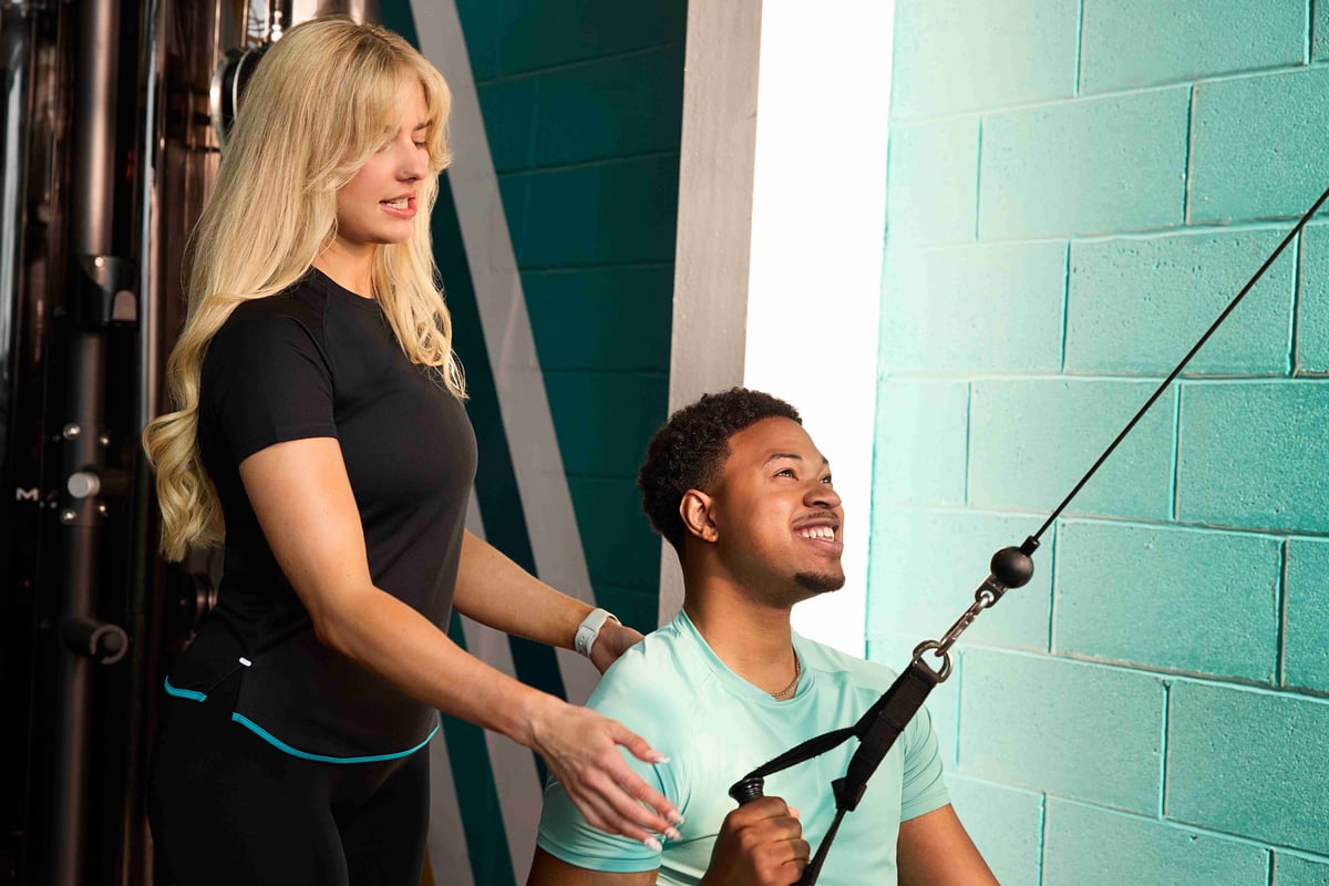 Woman coaching man using cable machine in gym with teal wall background