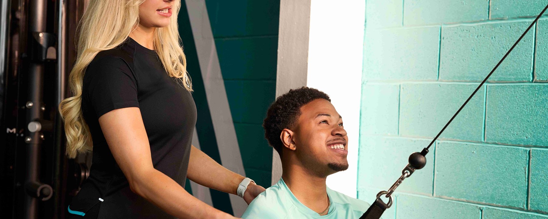 Woman coaching man using cable machine in gym with teal wall background