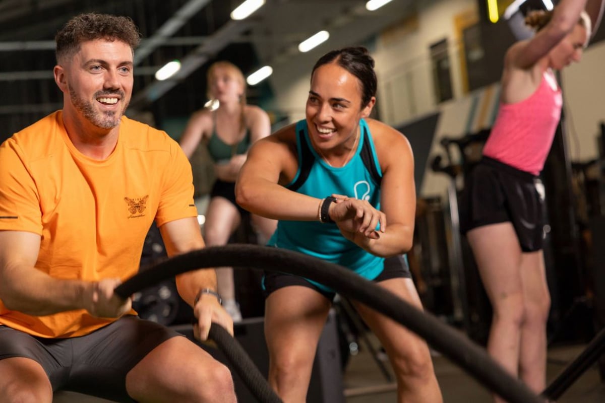 Man in orange shirt and woman in turquoise vest exercising on battle ropes in a gym