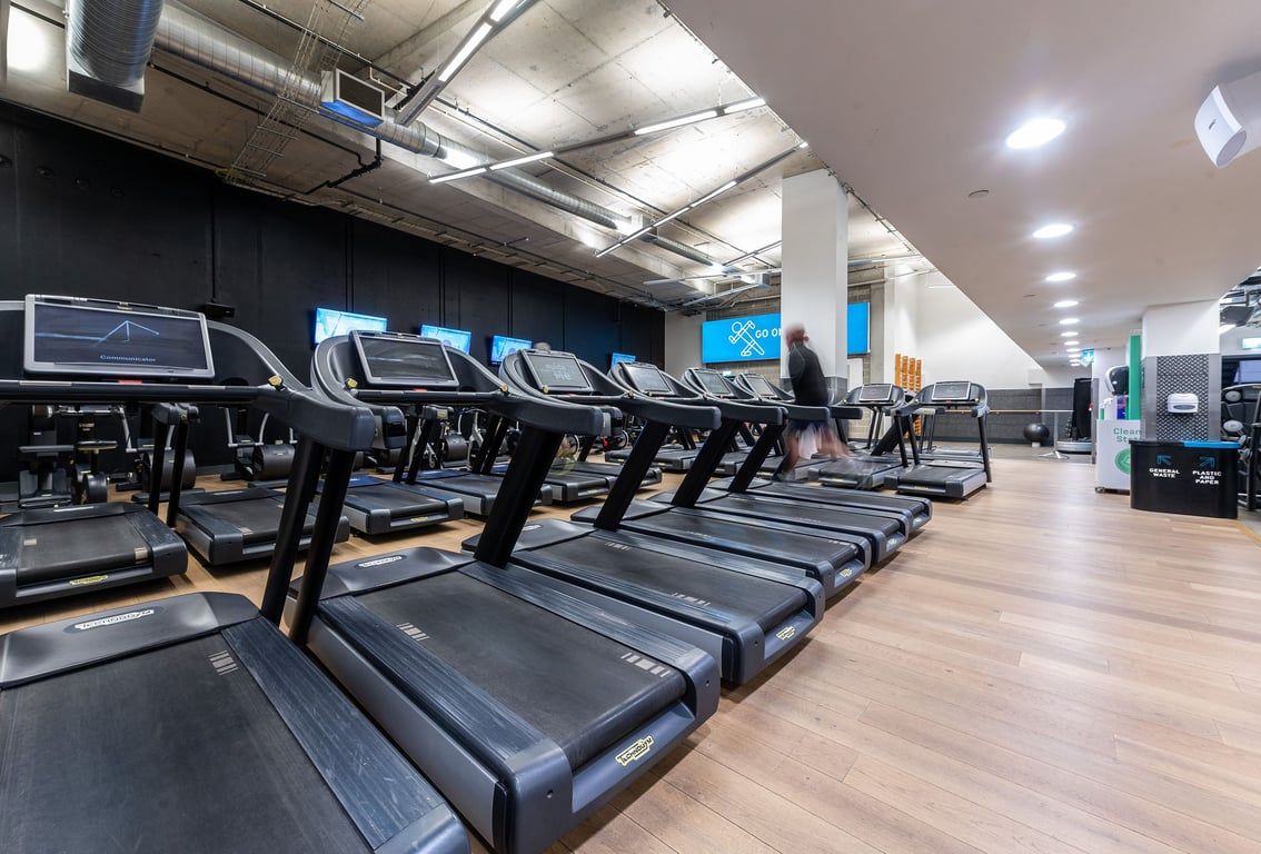 PureGym London Aldgate: Rows of treadmills inside a dimly lit gym space with industrial ceiling pipes.