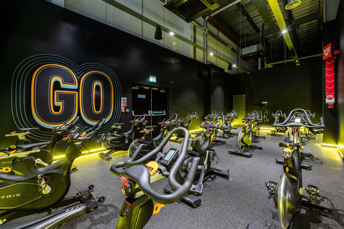 PureGym Peterborough Serpentine Green Shopping Centre: Rows of stationary bikes in a studio with neon "GO" signage.
