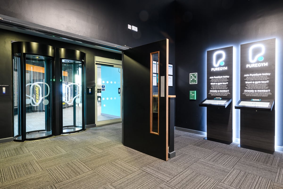 PureGym Leeds Thorpe Park: Row of sleek glass doors, illuminated signage and interactive kiosks - entrance area.