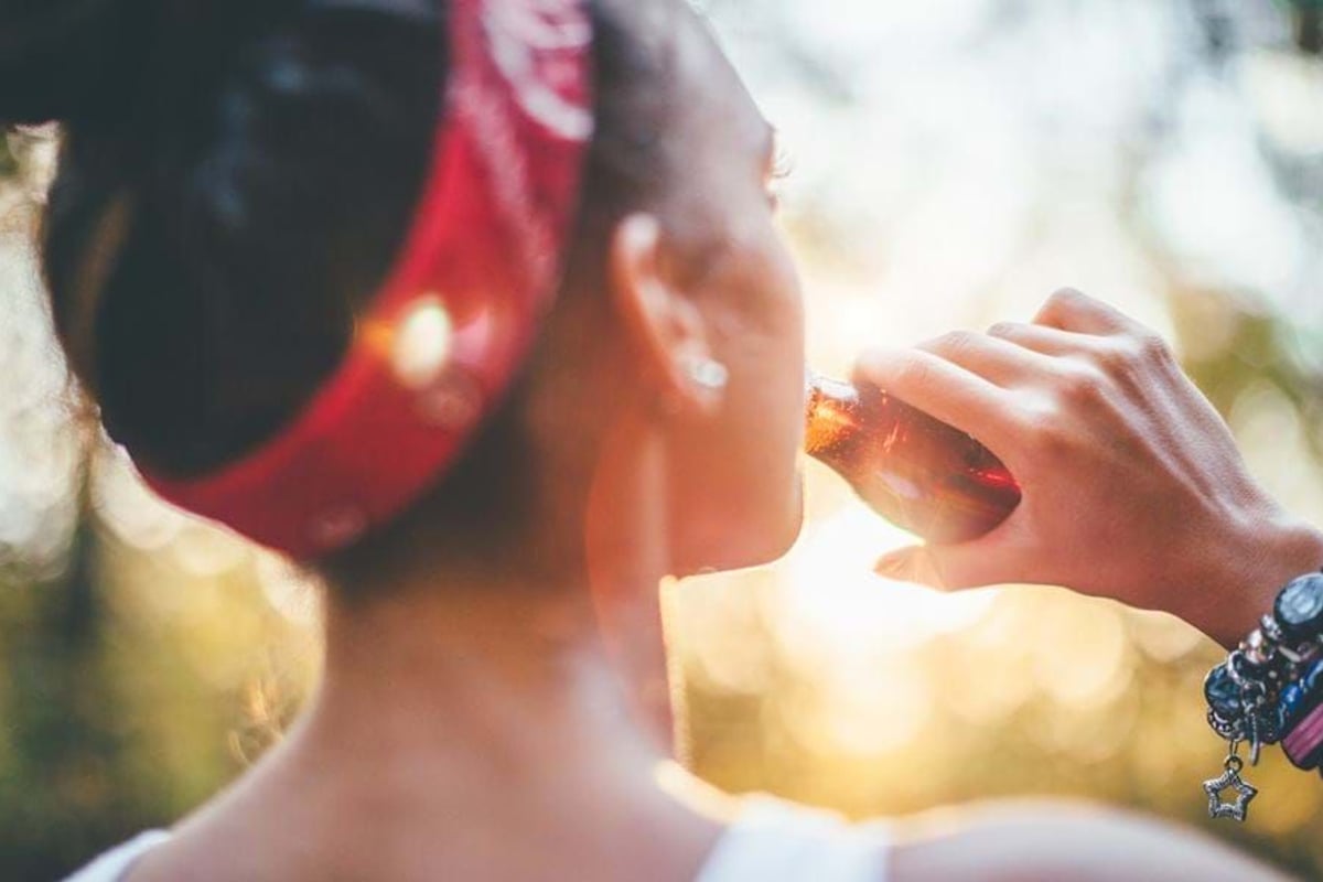 Woman with red headband and bracelets drinking from a brown glass bottle outdoors at sunse