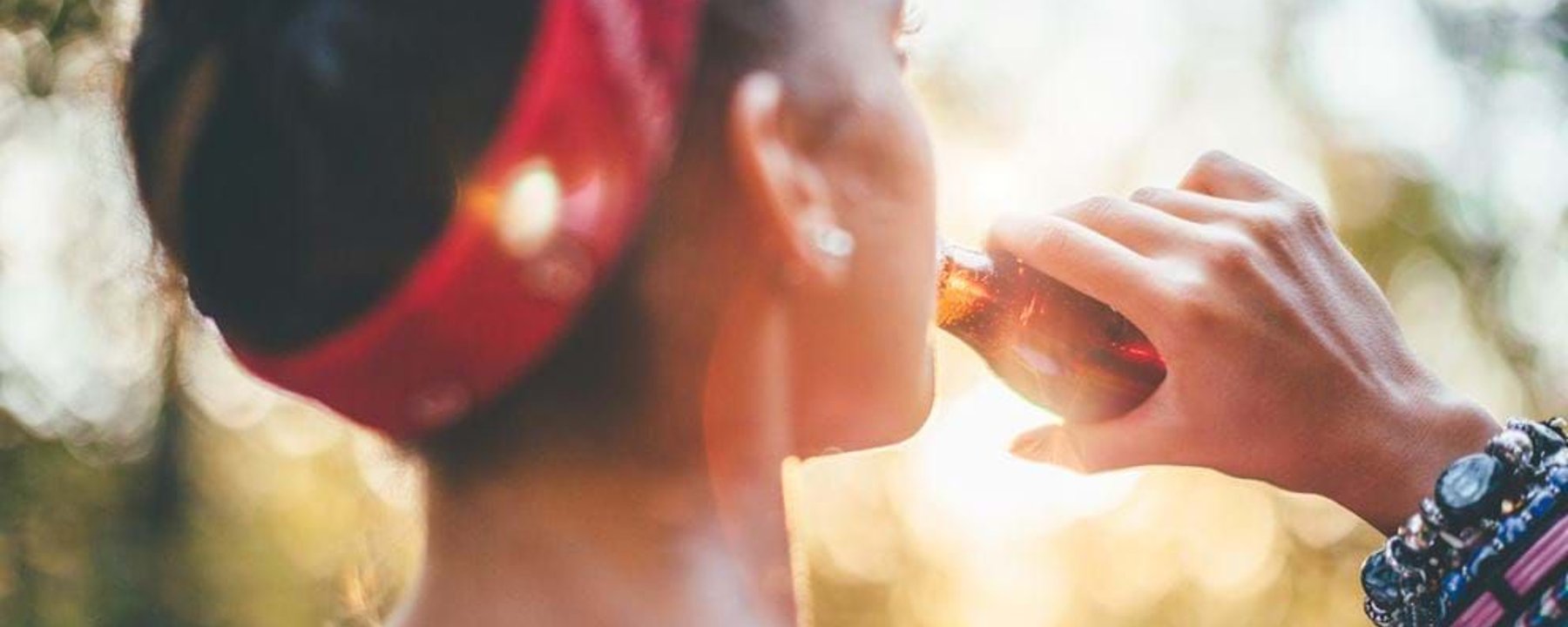 Woman with red headband and bracelets drinking from a brown glass bottle outdoors at sunse