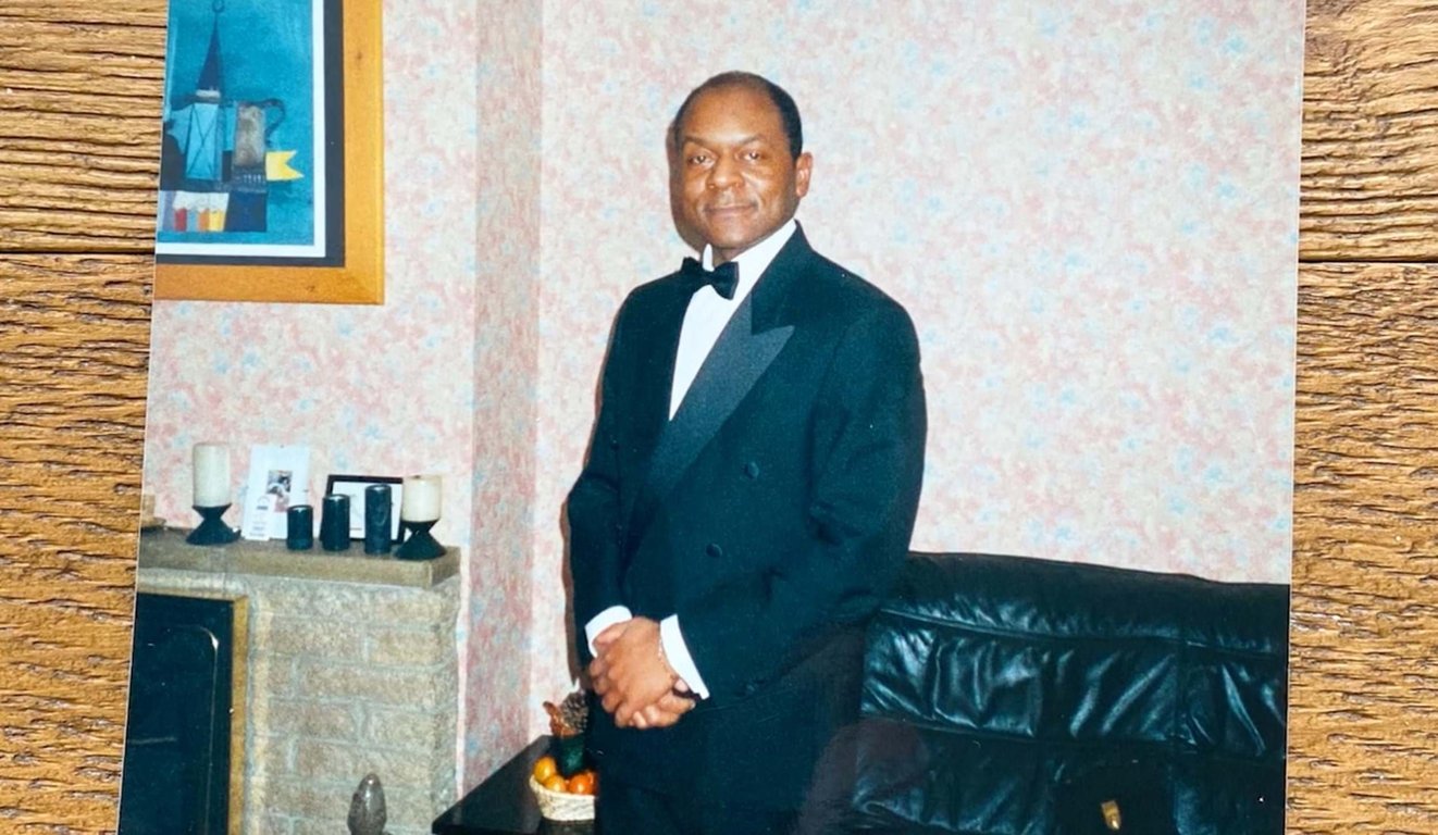 Man in black tuxedo standing in a living room with a leather sofa and framed art on wall