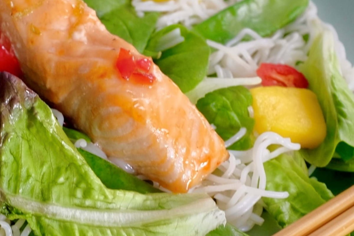 Grilled salmon on a bed of rice noodles, leafy greens, mango, and chopsticks on a plate