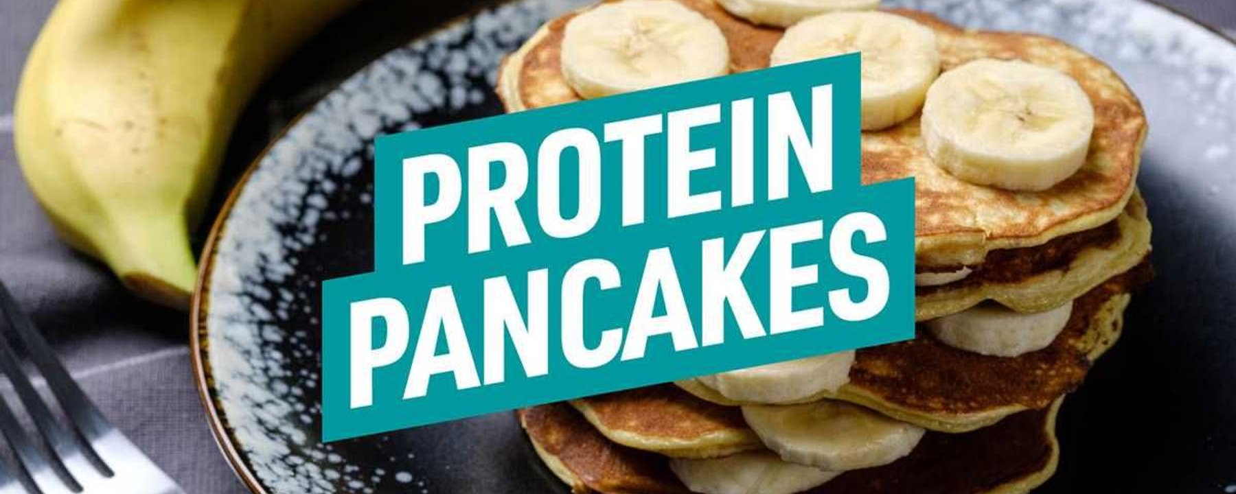Stack of banana slices topped pancakes on a black plate with bananas and fork nearby