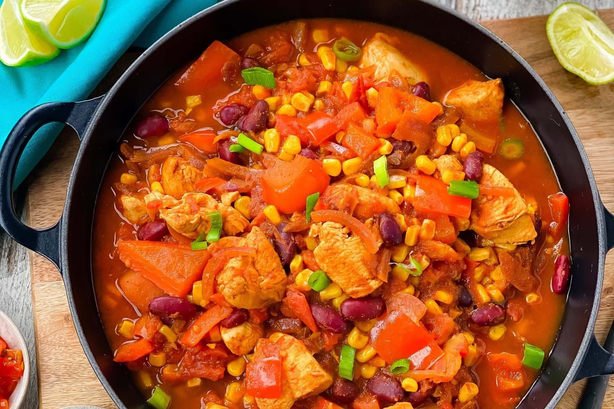 Chicken stew with corn, kidney beans, and red peppers in a black pan with rice and guacamole