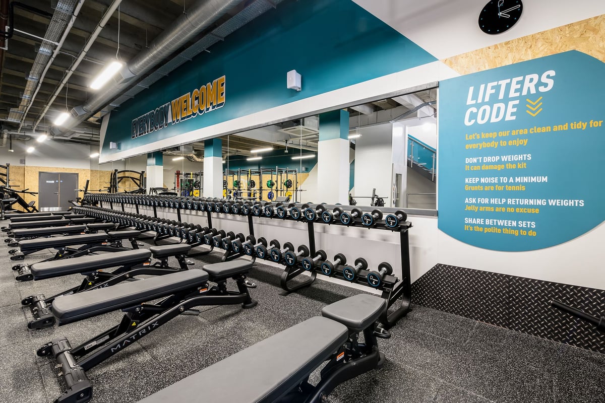 PureGym London Wimbledon: Free weight benches, dumbbell racks, rubberized flooring in a gym weightlifting area.