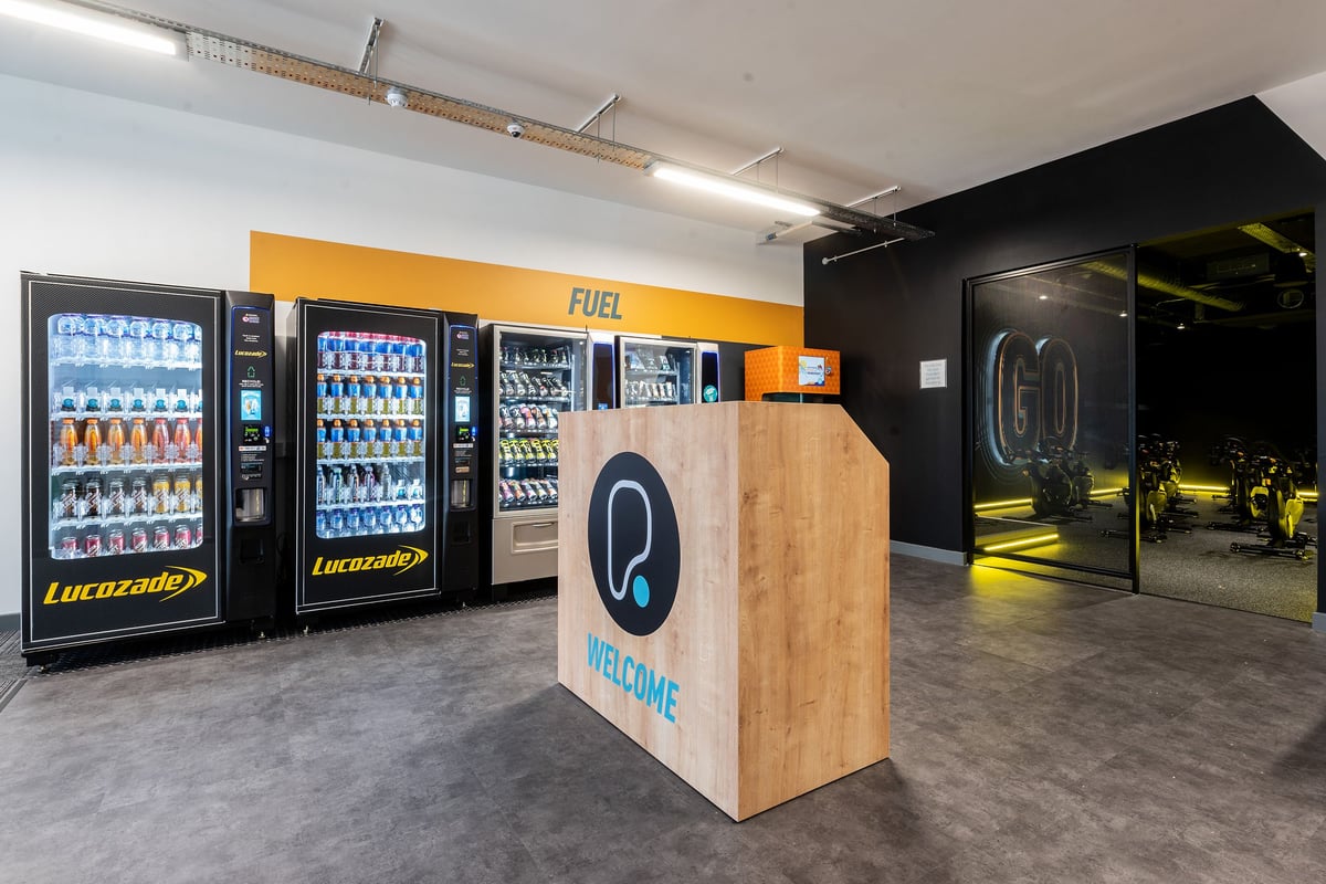 PureGym London Catford Rushey Green: Vending machines, a welcome sign, and entry gates in a modern fitness facility interior.
