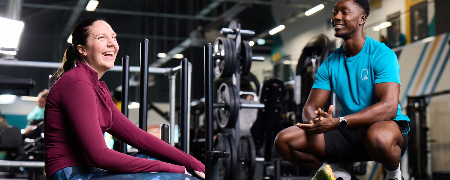 A man and woman sit on the gym floor, engaged in conversation, surrounded by exercise equipment.