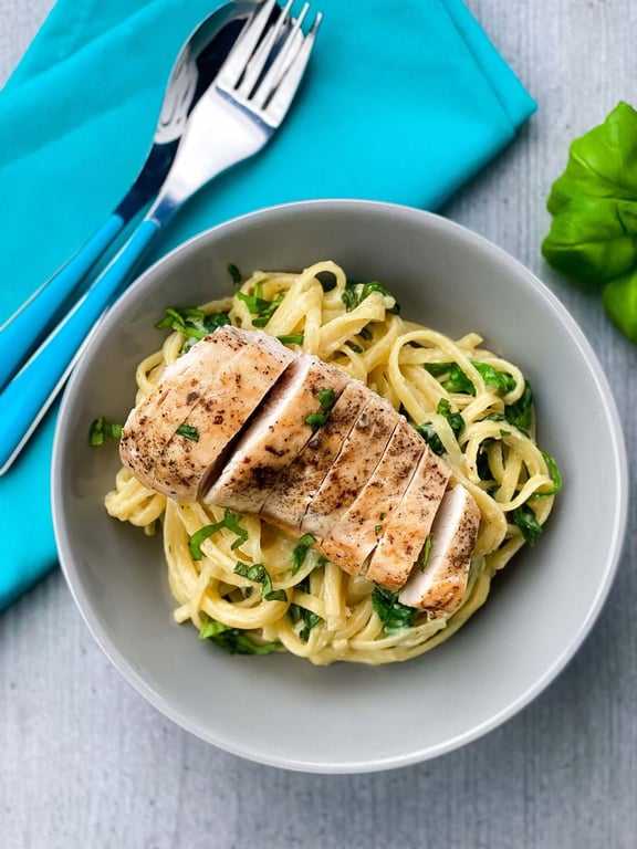 Grilled chicken breast slices on creamy pasta with spinach in a grey bowl with cutlery.