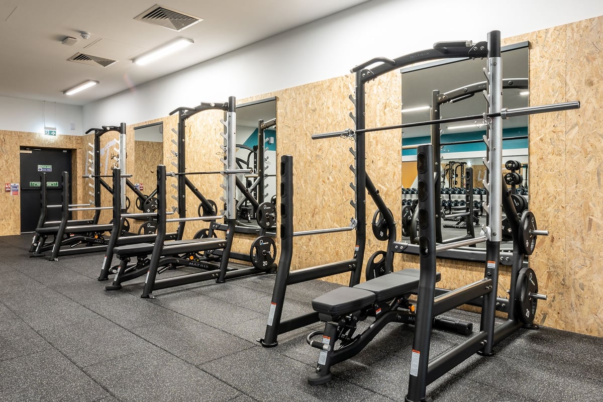 PureGym Glasgow Giffnock: Squat racks, benches, barbell plates, and wall-mounted exercise equipment on rubberized flooring.
