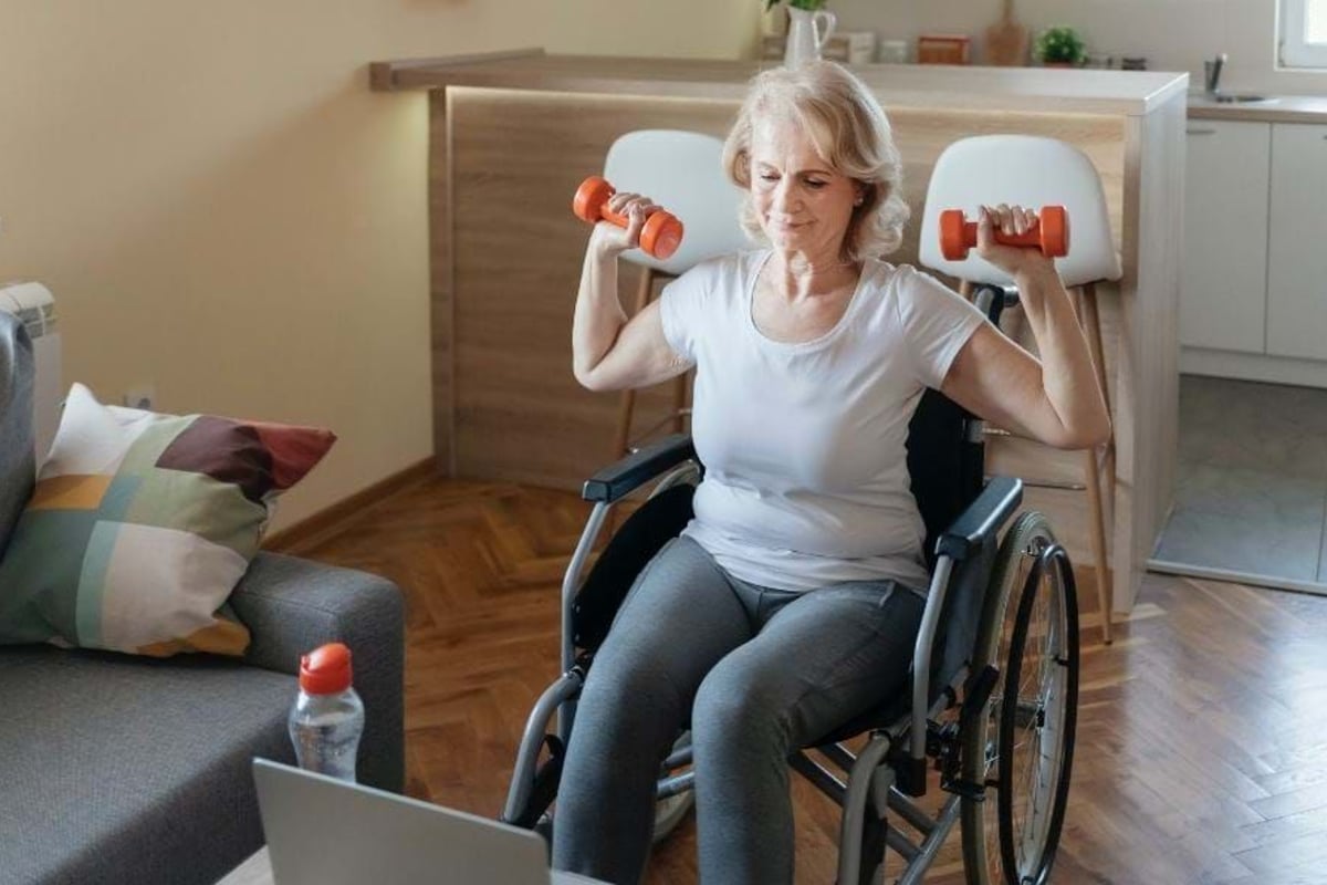 Elderly woman in wheelchair lifting dumbbells in living room near laptop.