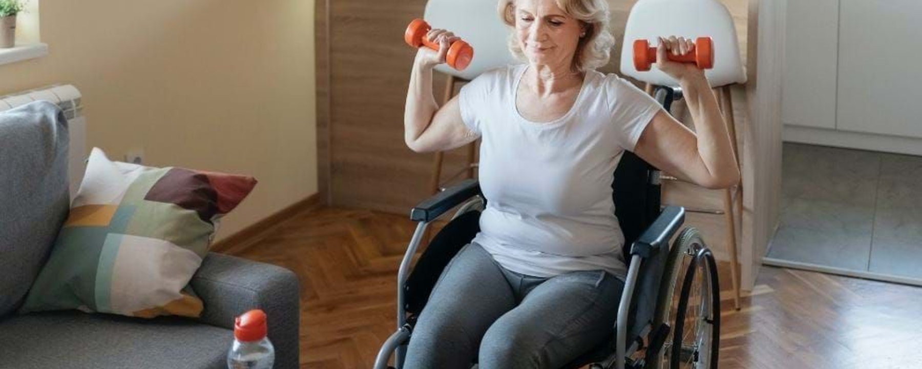 Elderly woman in wheelchair lifting dumbbells in living room near laptop.