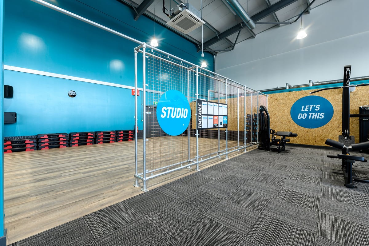 PureGym Maldon Blackwater Retail Park: Studio with teal walls, wire fencing, benches, weights, and rubberized flooring.