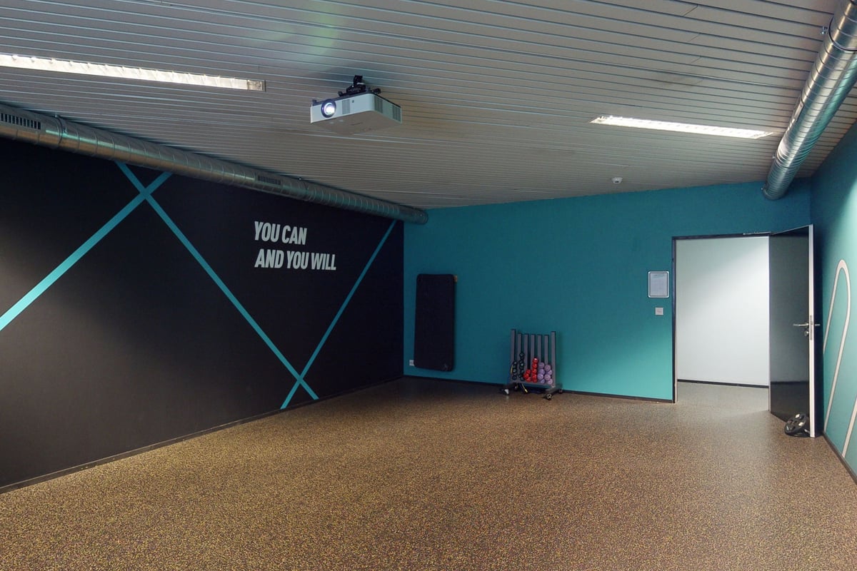 PureGym Kriens: Gym studio with teal walls, mirrored wall, free weights, rubberized flooring, and motivational text on wall.