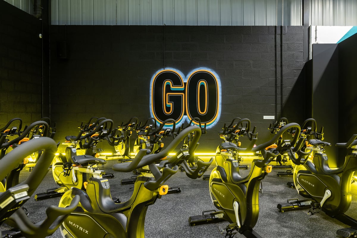 PureGym Dunfermline: Rows of yellow stationary exercise bikes, neon "GO" sign in a dimly lit space.