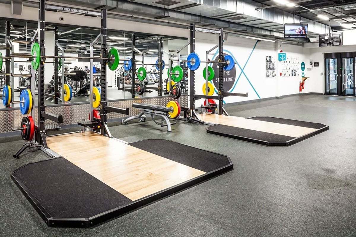 PureGym Altrincham: Weightlifting area with racks, platforms, wall graphics, and colourful resistance bands and balls.