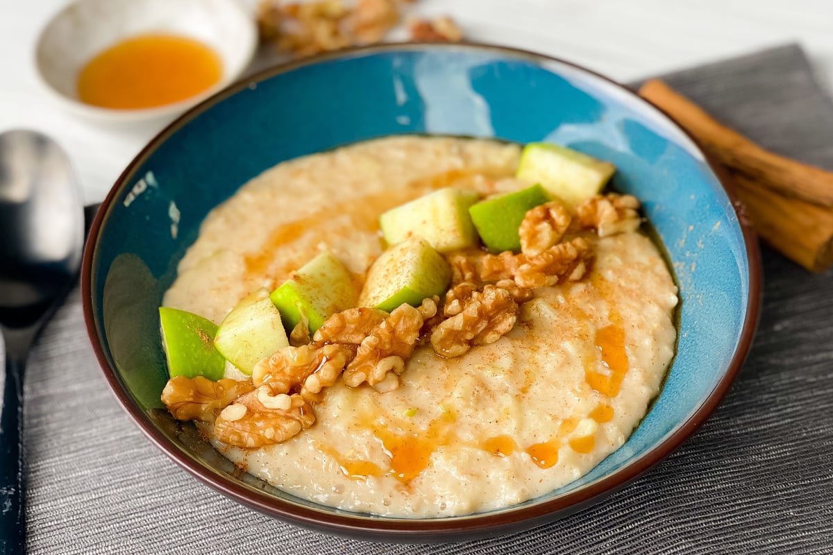 Bowl of porridge topped with chopped apple, walnuts and drizzle of honey on grey placemat