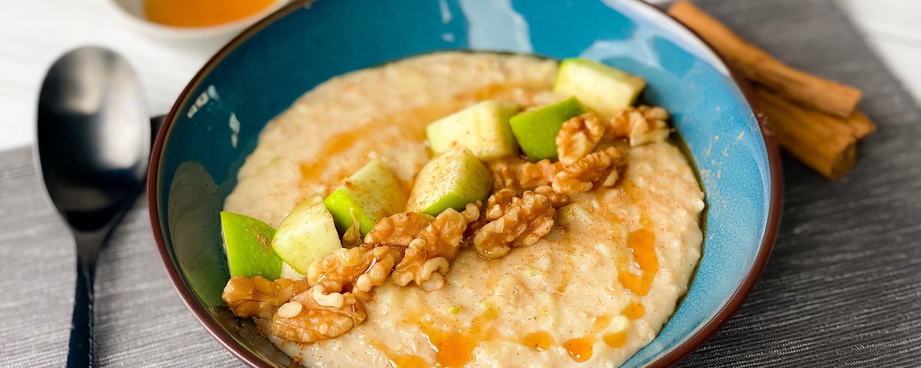 Bowl of porridge topped with chopped apple, walnuts and drizzle of honey on grey placemat