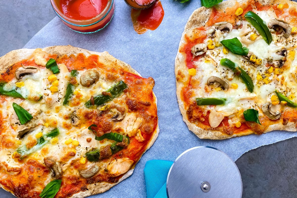 Two flatbread pizzas topped with chicken, peppers, mushrooms and sweetcorn on grey surface