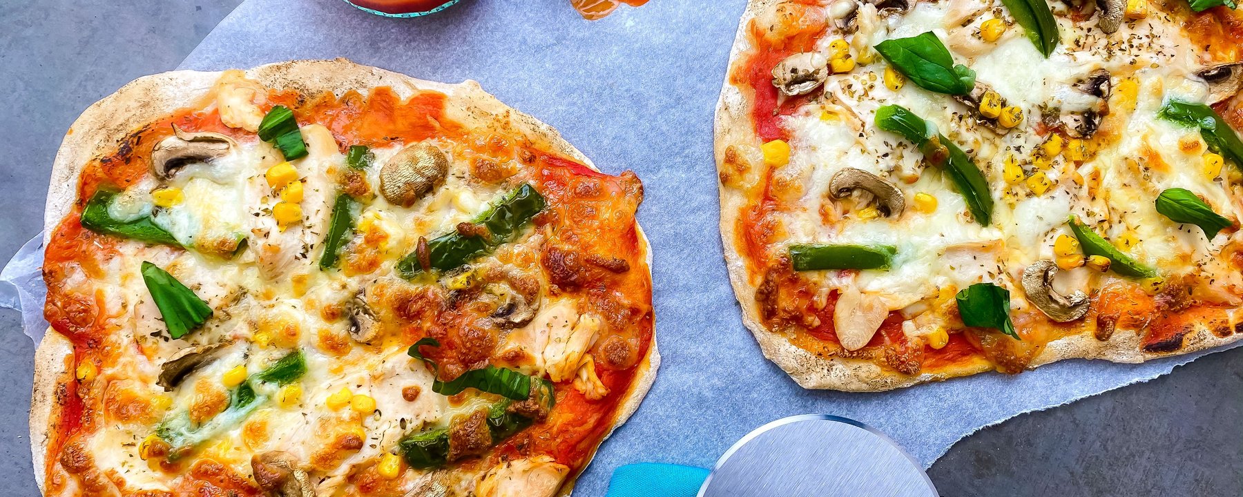 Two flatbread pizzas topped with chicken, peppers, mushrooms and sweetcorn on grey surface