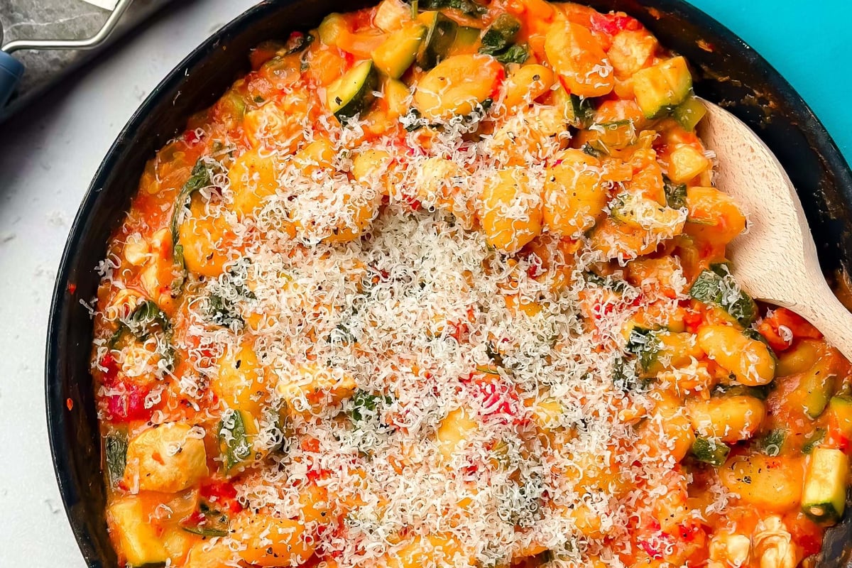 Pan of gnocchi with vegetables and grated cheese on top next to cheese grater and blue nap