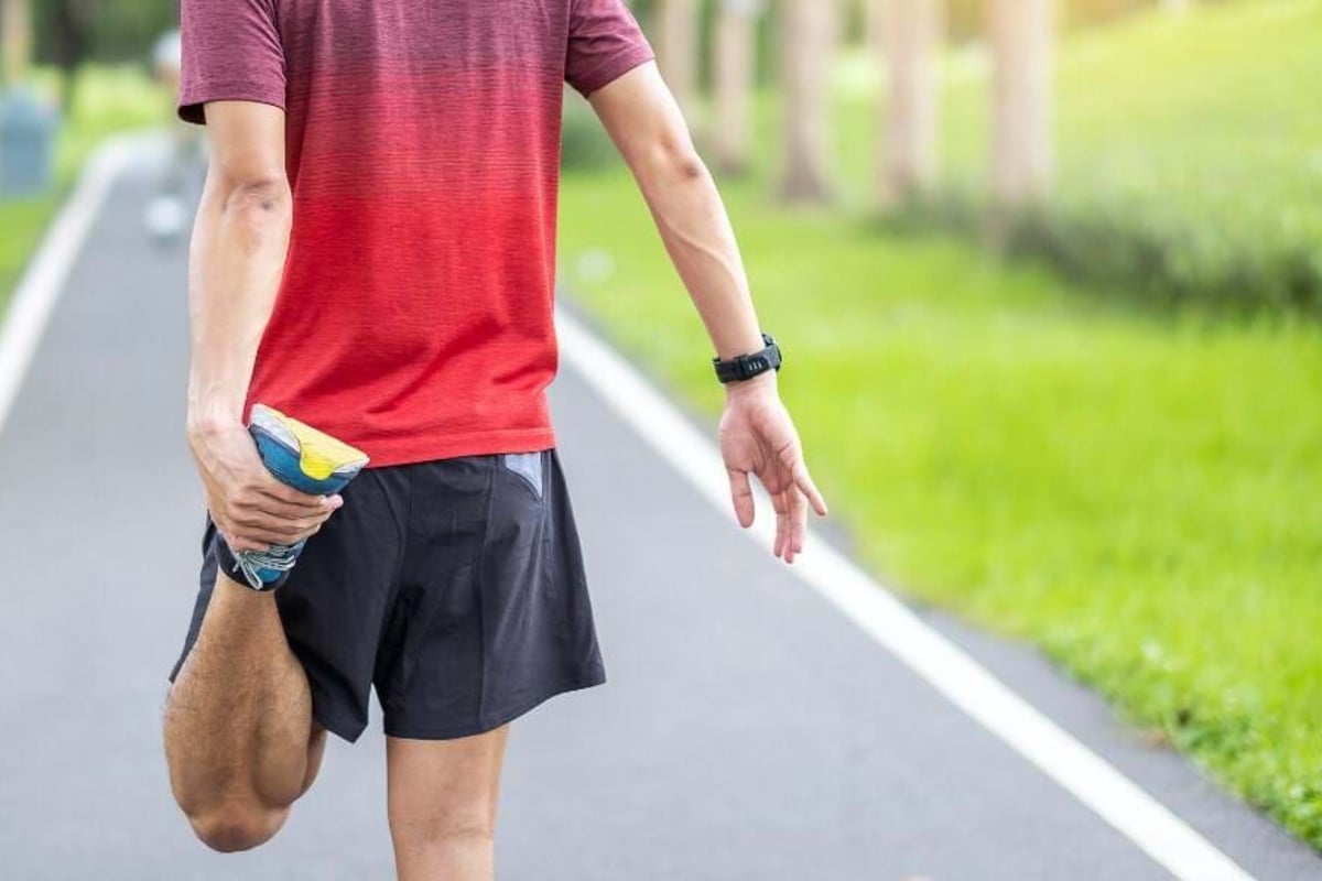 Man in red shirt stretching leg on a running track beside green grass.