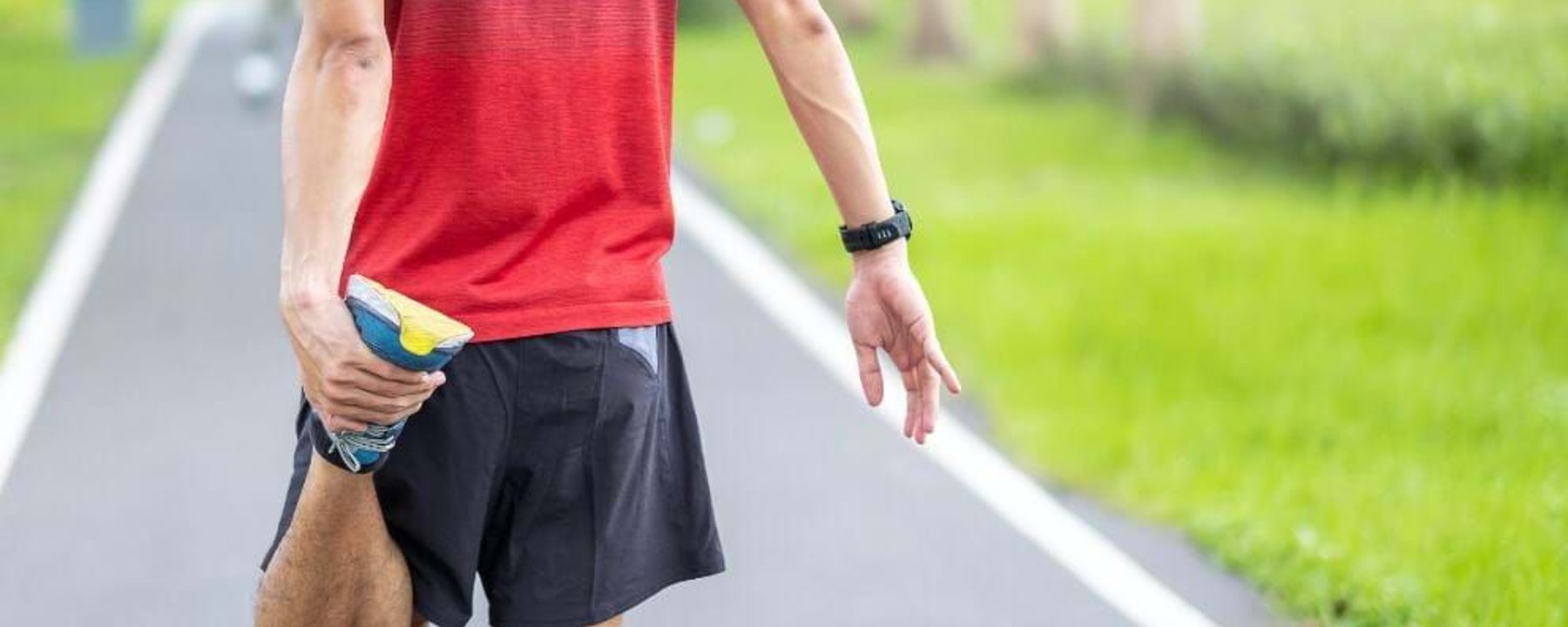 Man in red shirt stretching leg on a running track beside green grass.