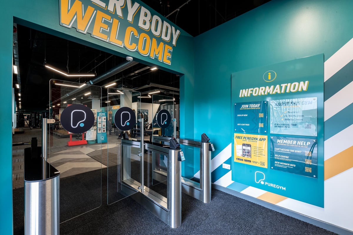 PureGym Hatfield: Entry turnstiles, welcome signage, blue and orange colour scheme, information board.