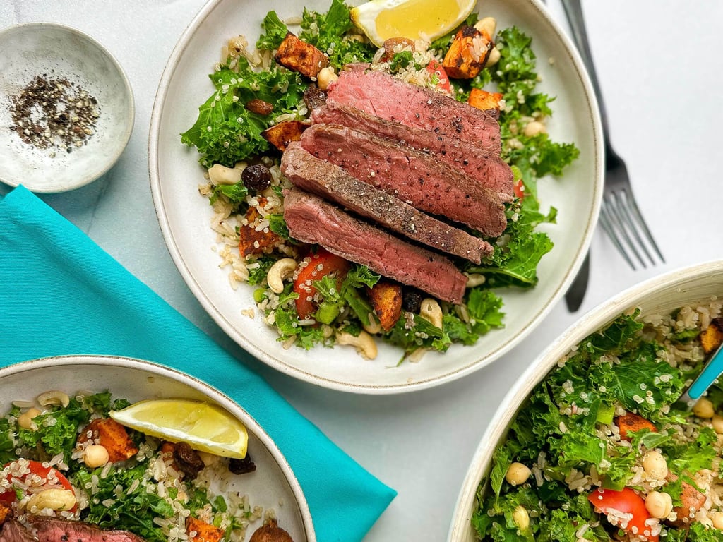 Sliced steak on a bed of kale, quinoa, nuts, and roasted vegetables with lemon wedges.