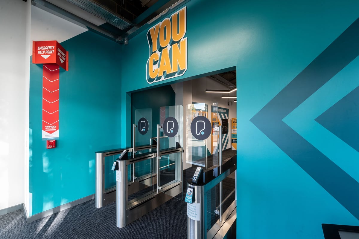 PureGym London Woolwich: Self-service kiosks, turnstiles, and a "You Can" sign in a turquoise-colored gym space.