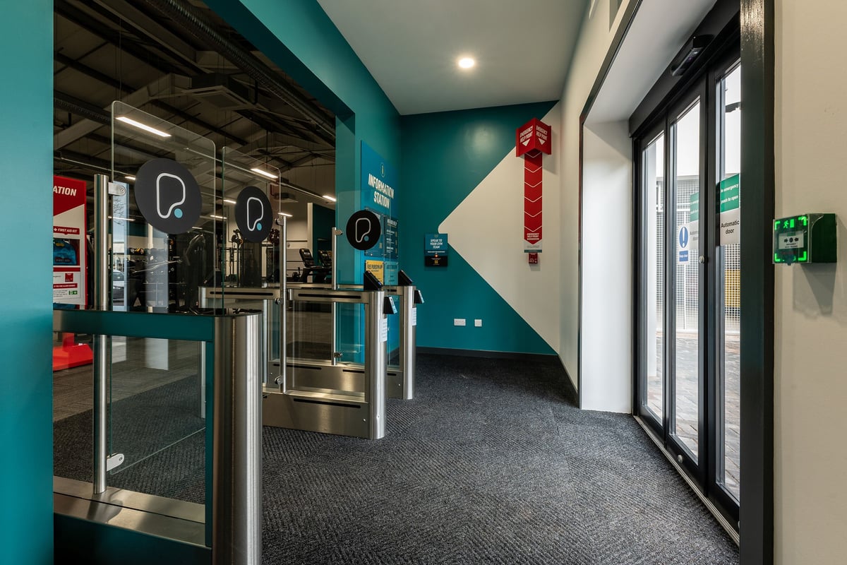 PureGym Uttoxeter: Turnstiles, lockers, and fire extinguishers in a teal-coloured hallway.
