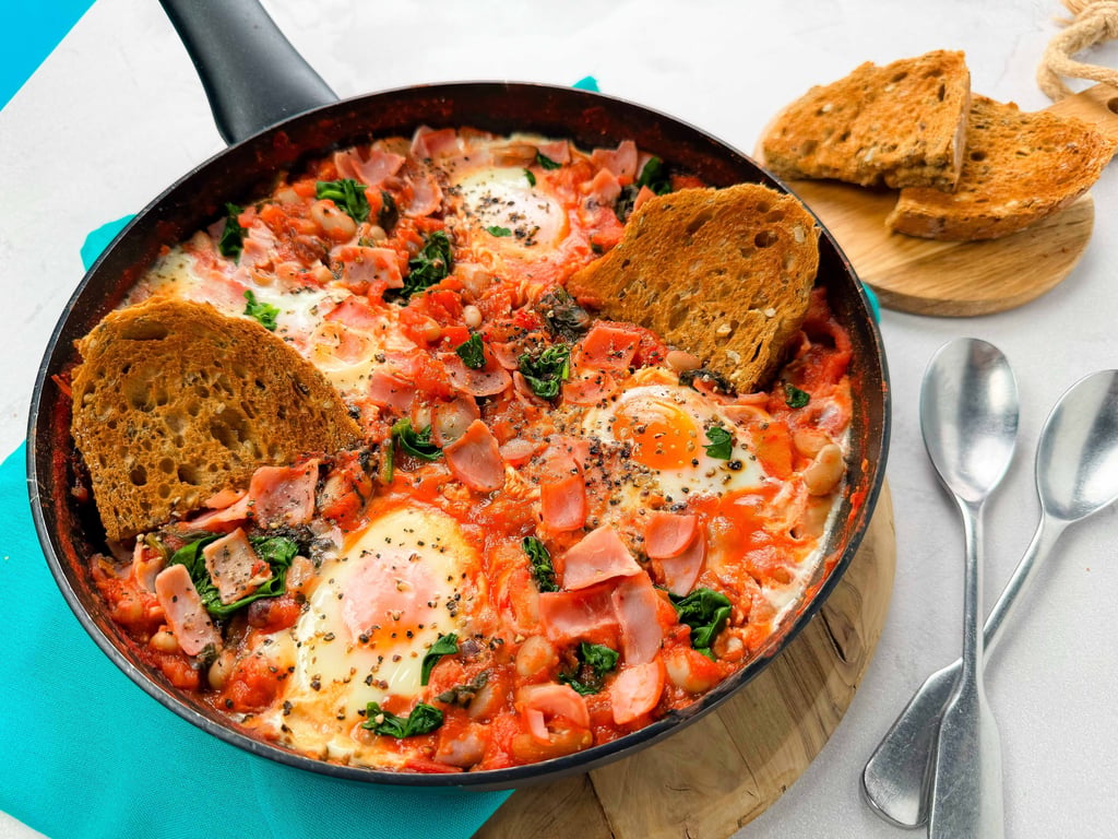 Shakshuka with eggs, spinach, ham, and toasted bread in a frying pan with spoons nearby