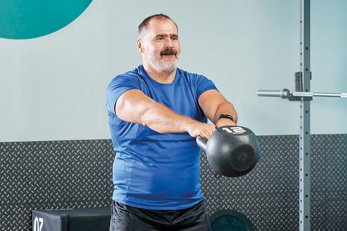 Man in blue shirt lifting a 12 kg kettlebell in a gym with weights in the background