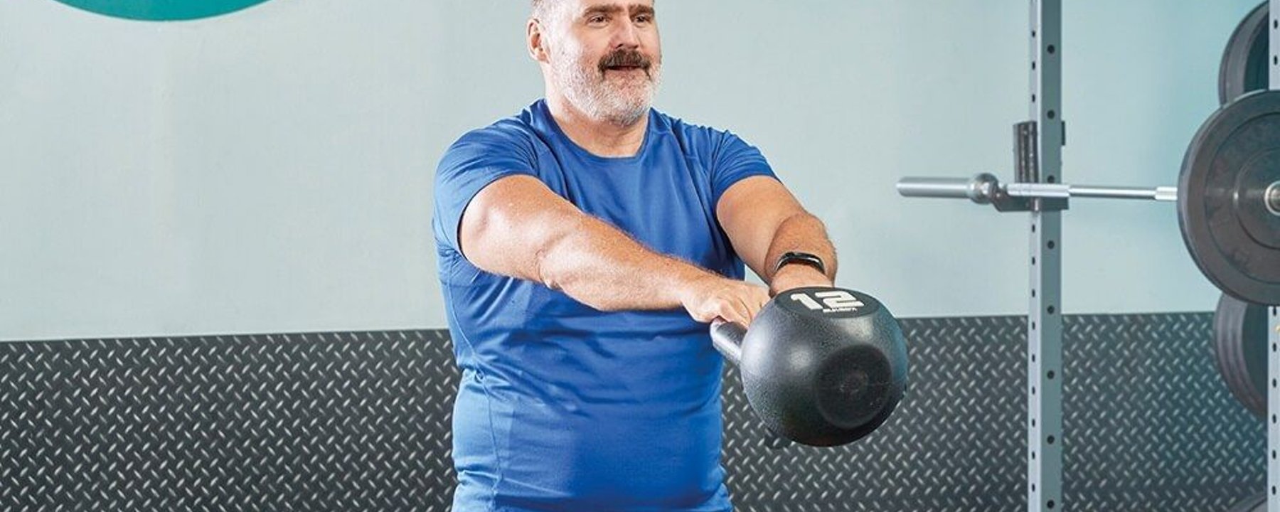 Man in blue shirt lifting a 12 kg kettlebell in a gym with weights in the background