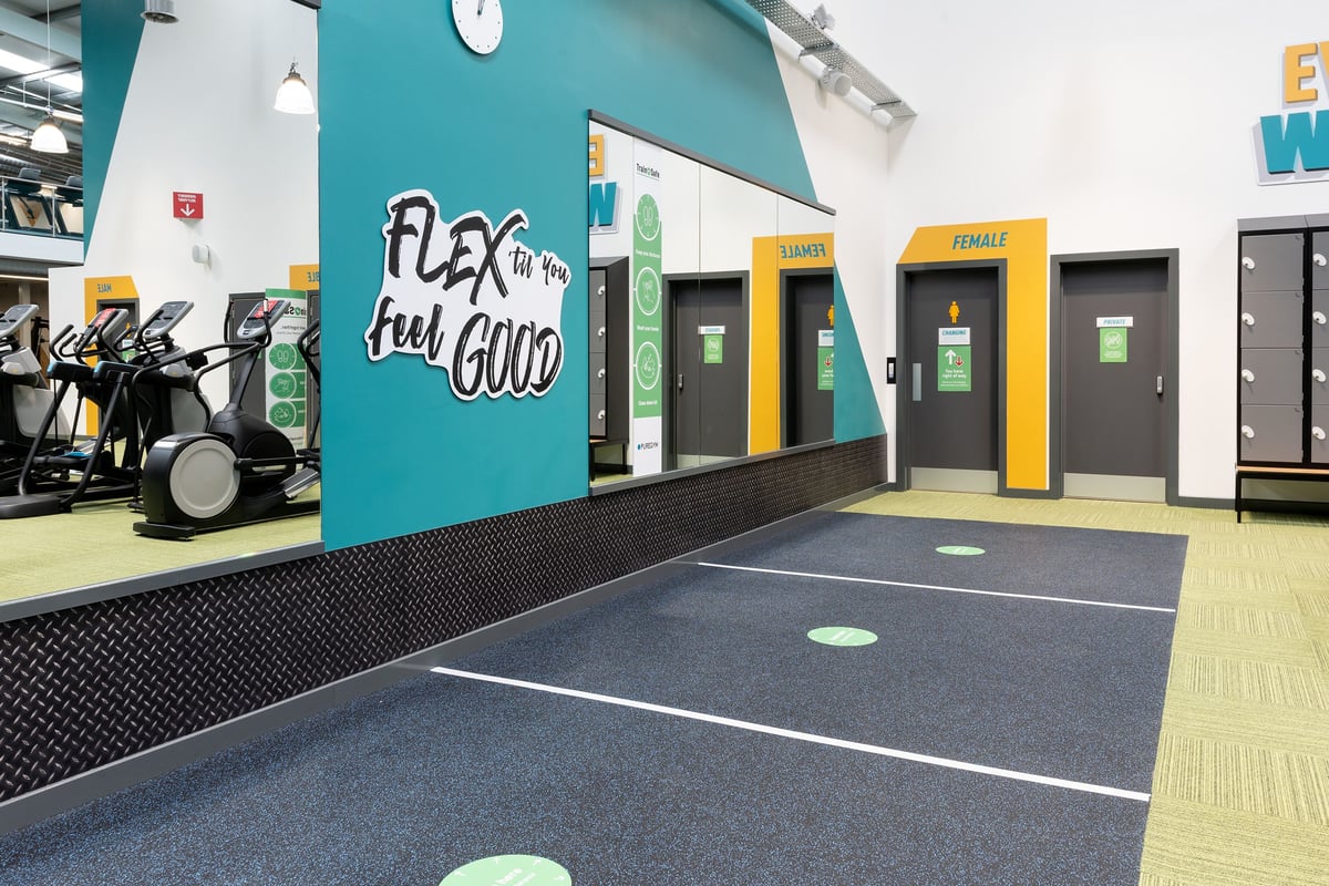PureGym Colchester Retail Park: Rows of exercise bikes, gym turf flooring, strength equipment, and colorful wall graphic.