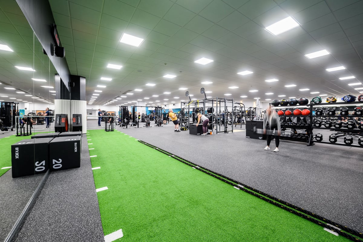 PureGym Aylesbury: Weight racks, free weights, green turf area, exercising people in a brightly lit gym interior.