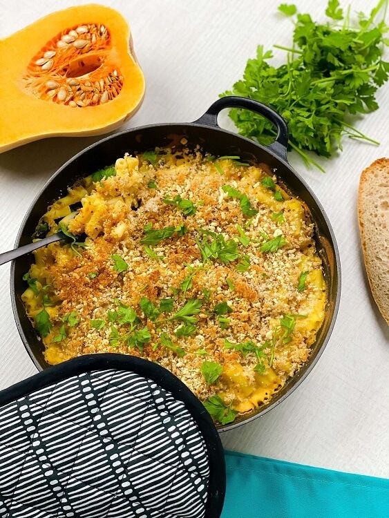 Baked macaroni and cheese with herb garnish in a black pan with pumpkin and bread nearby