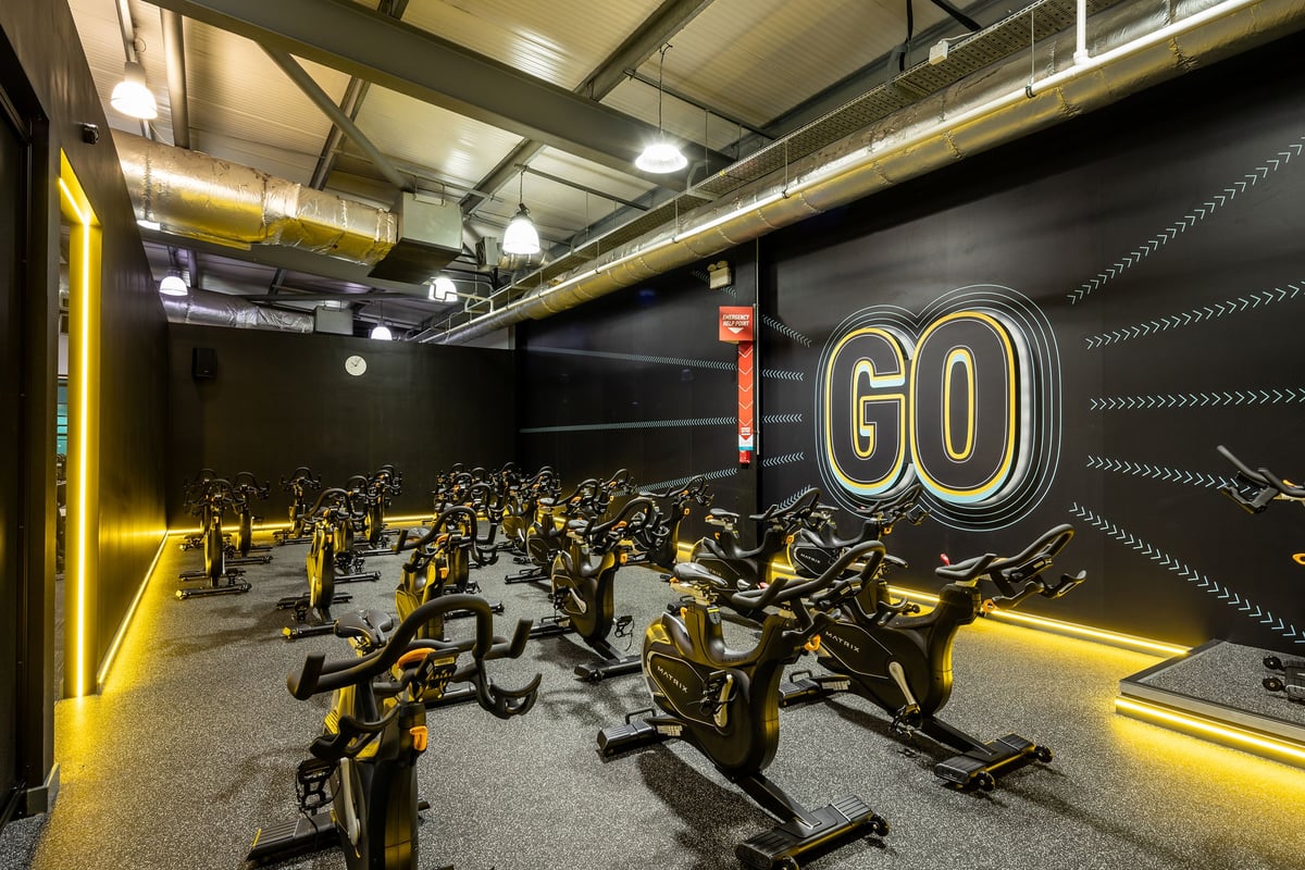 PureGym Cardiff Gate: Row of stationary cycles in a dimly lit studio with neon 'GO' sign and industrial ceiling.