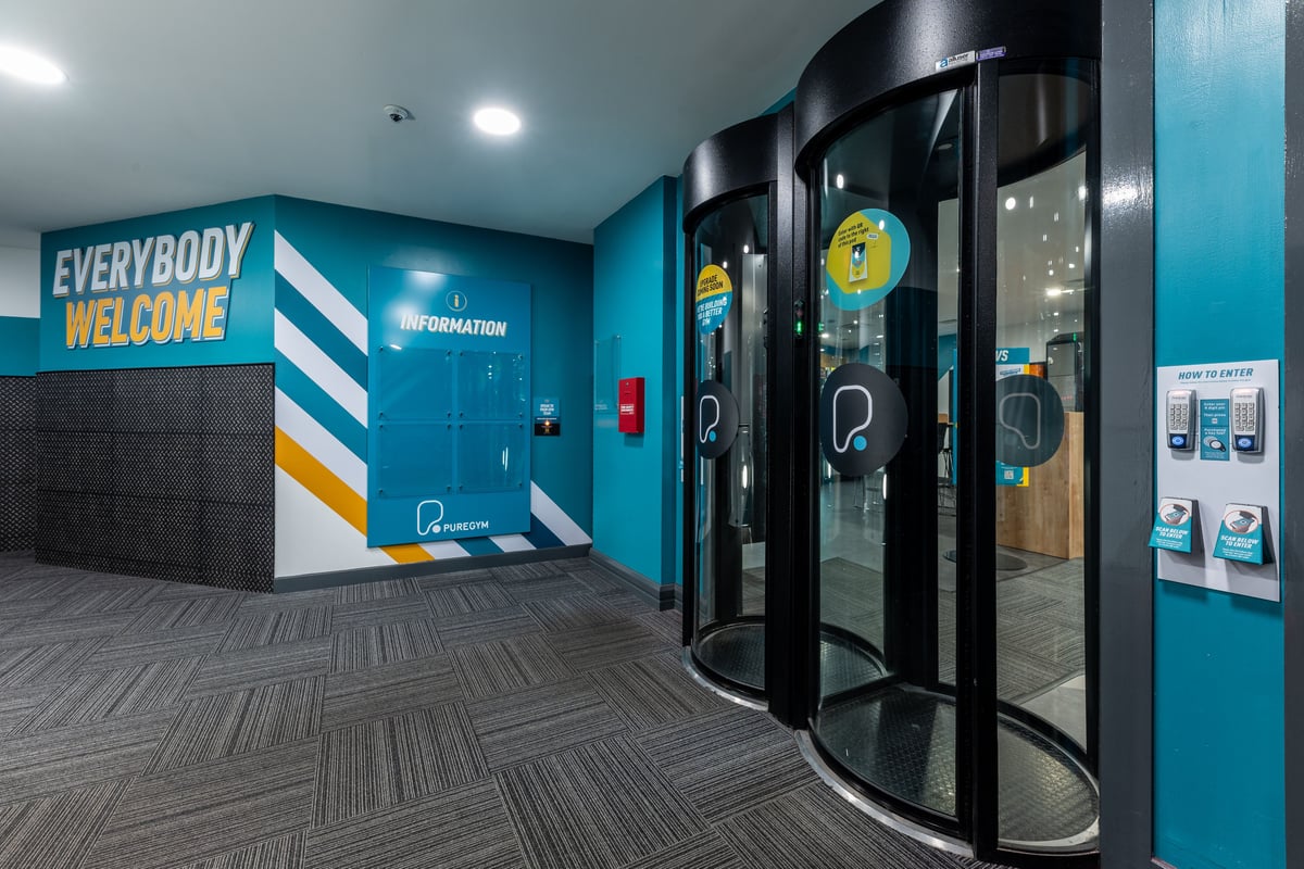 PureGym London Edgware: Entrance area with information kiosks, revolving gates, teal walls, and "Everybody Welcome" signage.